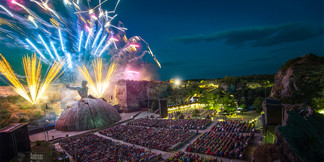 Open-Air-Theater in Felsenlandschaft bei Nacht mit Feuerwerk über der Bühne und großem Publikum.
