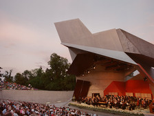 Blick auf den modernen Wolkenturm in Grafenegg bei Sonnenuntergang, mit einem Orchester auf der Bühne und zahlreichen Zuschauer:innen im Freiluftbereich.