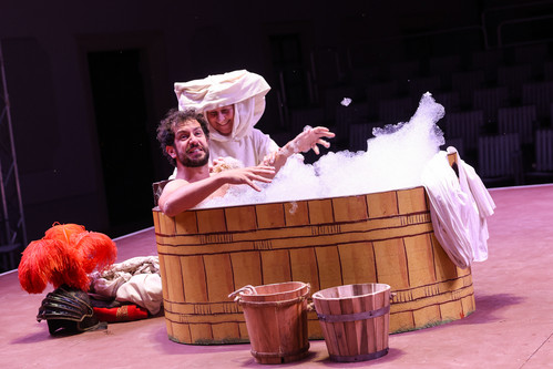 Theaterszene: Zwei Darsteller:innen sitzen lachend in einer überdimensionalen, mit Schaum gefüllten Holzzuber; auf dem Boden davor zwei hölzerne Wassereimer und ein roter Federhelm.