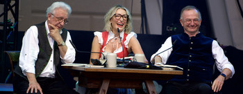 Drei Personen in Tracht sitzen lachend an einem Holztisch mit Mikrofonen und Bierkrügen auf einer Bühne.