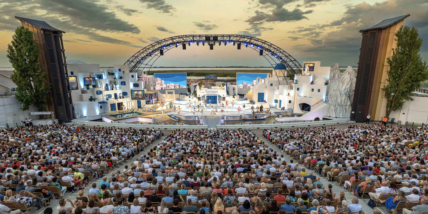 Großes Open-Air-Theater mit vollbesetztem Publikum vor mediterraner Bühnenkulisse bei Sonnenuntergang.