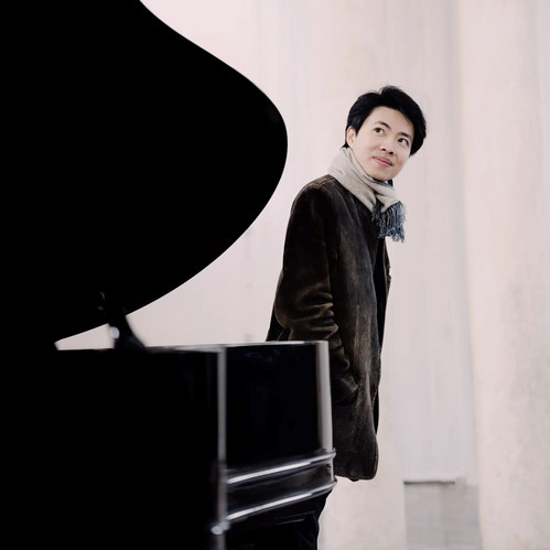 Kit Armstrong steht neben einem Konzertflügel in heller Umgebung, im dunklen Mantel mit Schal – nachdenklicher Blick zur Seite, moderner klassischer Pianist.