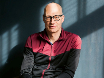 Schauspieler Joachim Meyerhoff in sportlicher Jacke mit verschränkten Händen auf einem Holzstuhl sitzend, vor einer blauen Wand im Streiflicht.