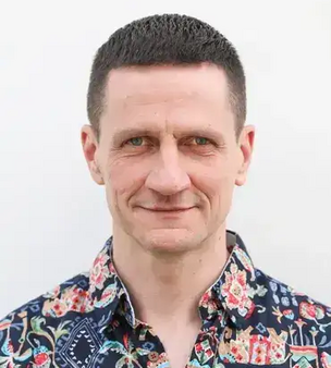 Porträt eines Mannes mit kurz geschnittenem dunklem Haar und gemusterter Hemdbluse vor weißem Hintergrund, mit direktem Blick und leichtem Lächeln.