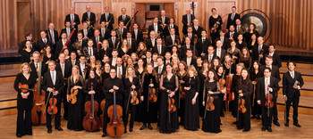 Das ORF Radio-Symphonieorchester Wien versammelt sich für ein offizielles Gruppenfoto auf einer Bühne mit Holzverkleidung; die Musiker:innen tragen elegante Konzertkleidung und halten ihre Instrumente.