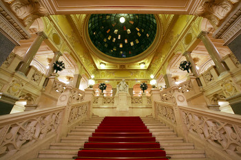 Prunkvolles Treppenhaus mit rotem Teppich, Statuen und verziertem Glasdach.