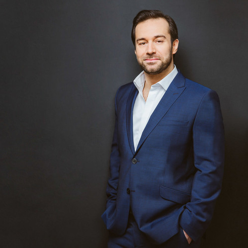 Mann im blauen Anzug steht lächelnd vor dunklem Hintergrund.