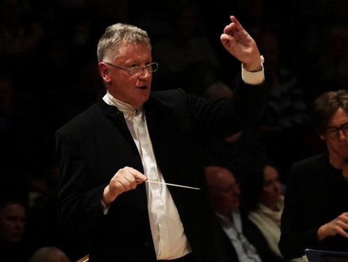 Dirigent in dunklem Anzug mit erhobener Hand und Taktstock vor Publikum.