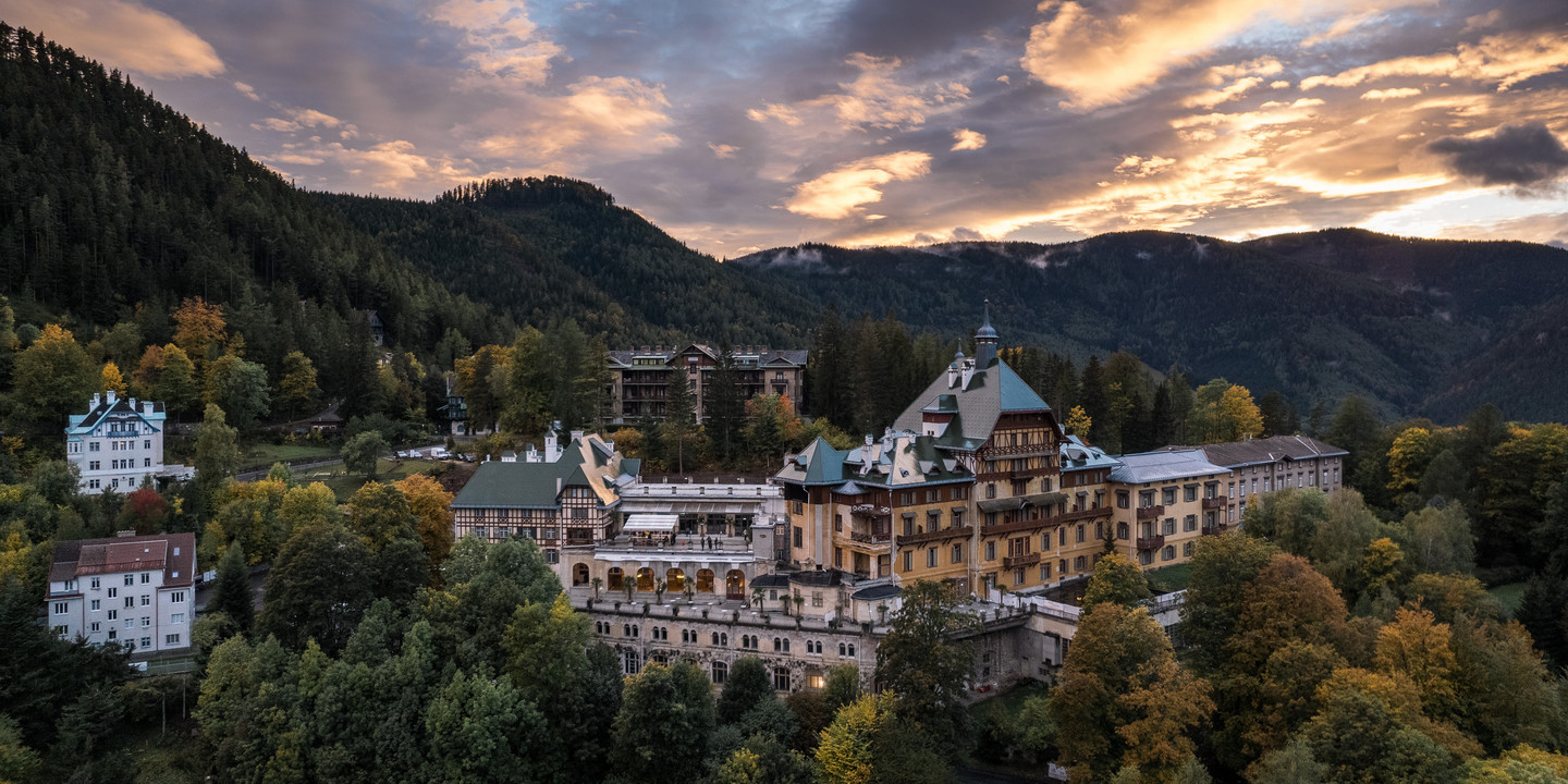 Historisches Grandhotel inmitten eines herbstlich gefärbten Waldes am Semmering, aufgenommen bei Sonnenuntergang mit dramatisch gefärbtem Himmel im Hintergrund.