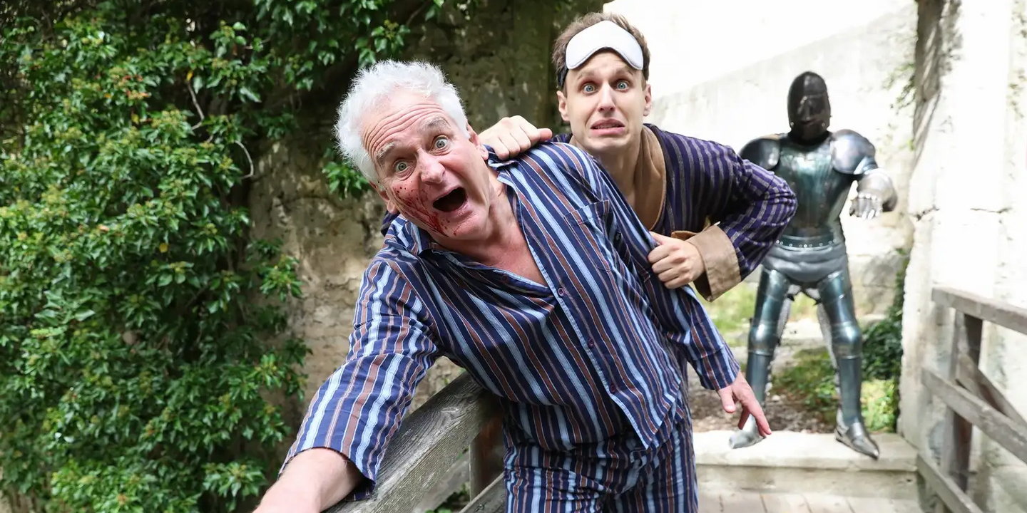 Zwei Männer im Pyjama auf einer Holzbrücke, der vordere blutverschmiert und mit entsetztem Gesichtsausdruck, der hintere mit Schlafmaske auf der Stirn und ängstlichem Blick; im Hintergrund eine Ritterrüstung – humorvolle Theaterszene in Freiluftkulisse.