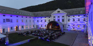 Open-Air-Bühne im Innenhof mit Publikum und bunter Fassadenbeleuchtung bei Abenddämmerung.