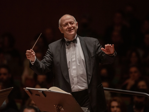 Dirigent Iván Fischer in schwarzem Frack mit erhobenen Armen beim Dirigieren, im Hintergrund unscharf das Publikum.