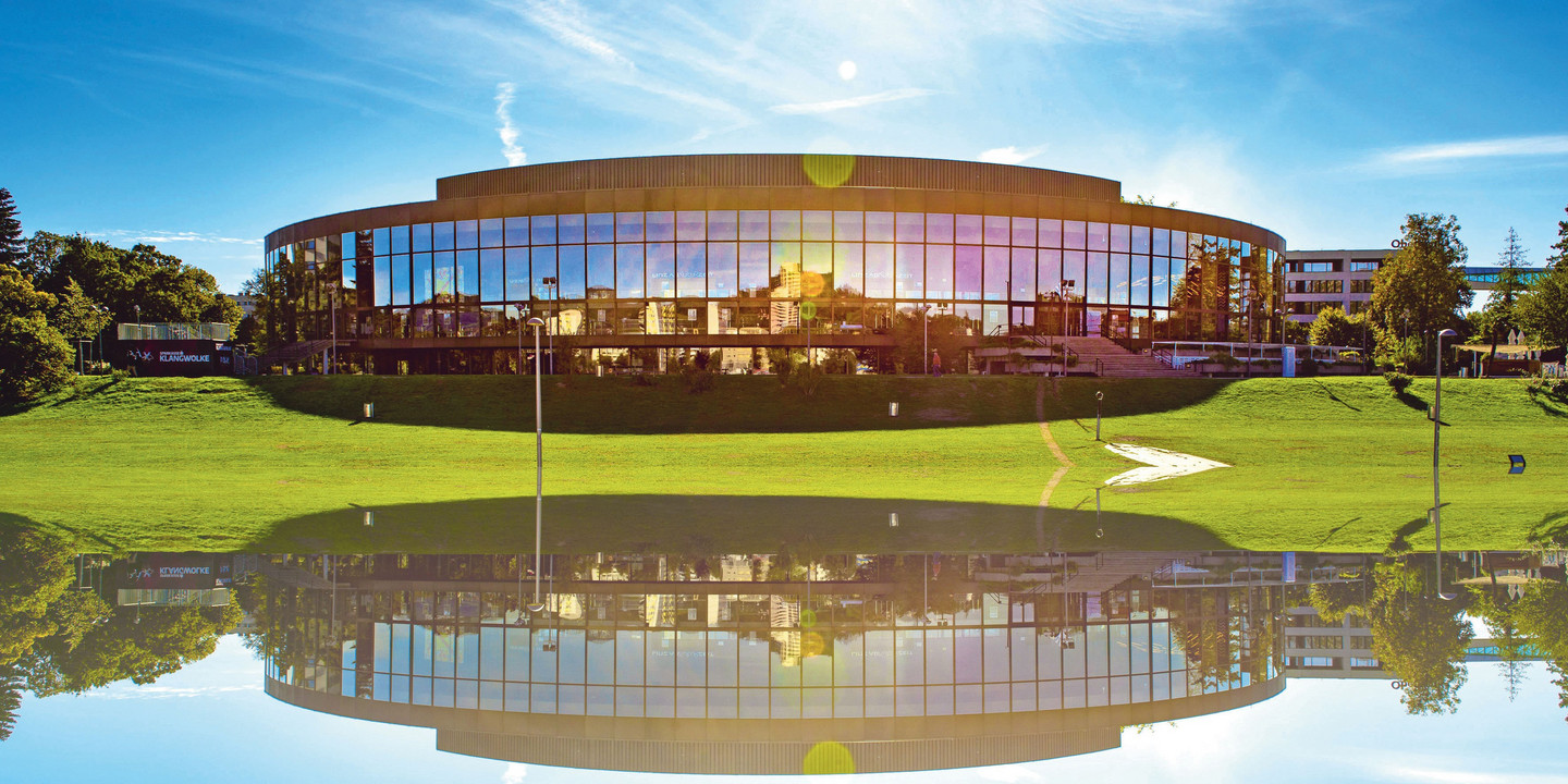 Modernes Konzerthaus mit Glasfassade, davor grüne Wiese und Wasserreflexion.