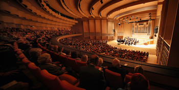 Moderner Konzertsaal mit Publikum und Orchester auf beleuchteter Bühne.