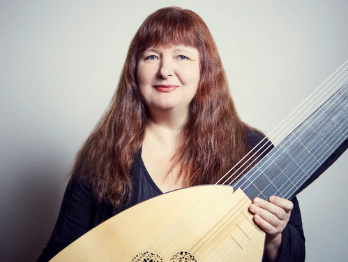 Frau mit langen rotbraunen Haaren in schwarzer Kleidung, die eine Laute vor neutralem Hintergrund hält.