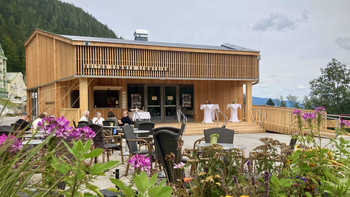 Moderner Holzpavillon des Kultur.Sommer.Semmering mit Terrasse, Gästen und Blumen im Vordergrund.