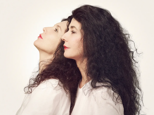 Künstlerporträt der Pianistinnen Katia und Marielle Labèque, im Profil Rücken an Rücken, mit weißen Oberteilen vor hellem Hintergrund.