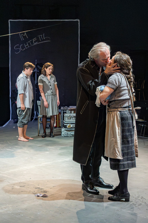 Intensive Bühnenszene: Ein älterer Mann küsst zärtlich eine Frau, während zwei jüngere Darsteller:innen im Hintergrund mit ernster Miene zuschauen. Das Bühnenbild ist minimalistisch, mit angedeuteter Wandinschrift „IM SCHETTL“ und gestapeltem Gepäck.