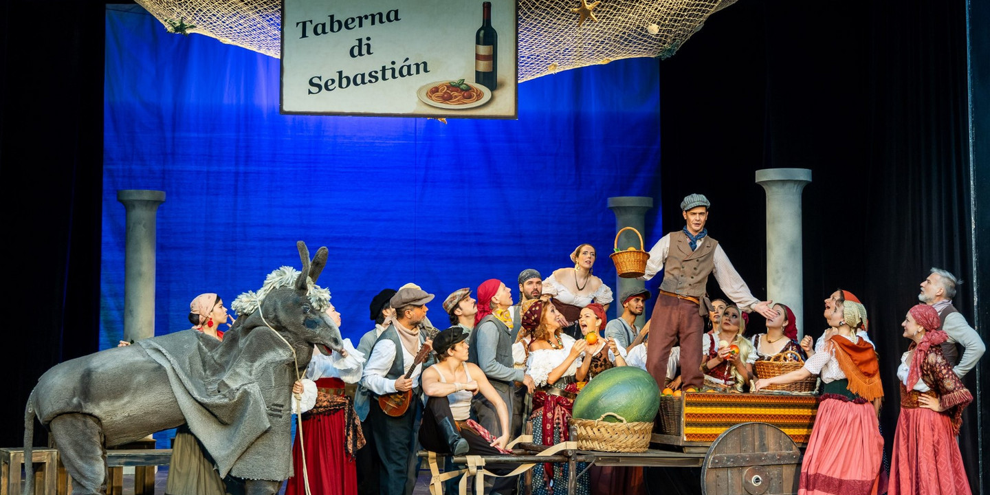 Bühnenbild einer italienisch inspirierten Theaterszene mit vielen Darsteller:innen in bunten, historischen Kostümen vor einem blauen Hintergrund. 