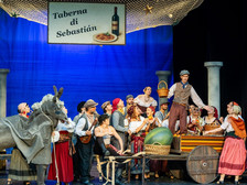 Bühnenbild einer italienisch inspirierten Theaterszene mit vielen Darsteller:innen in bunten, historischen Kostümen vor einem blauen Hintergrund. 