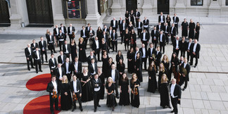 Großes Orchester in eleganter Kleidung posiert im Freien auf rotem Teppich vor historischem Gebäude.