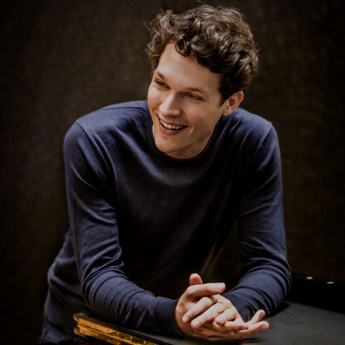 Lukas Sternath lehnt sich lächelnd an einen Konzertflügel, in lockerem Pullover, in entspannter und konzentrierter Pose – Porträt des österreichischen Pianisten.