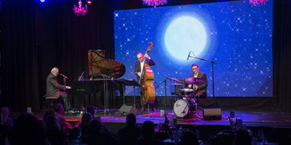 Jazz-Trio mit Klavier, Kontrabass und Schlagzeug vor nächtlichem Bühnenbild mit Mondprojektion