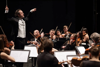 Dirigent mit erhobenen Armen leitet ein konzentriertes Orchester auf dunkler Bühne.