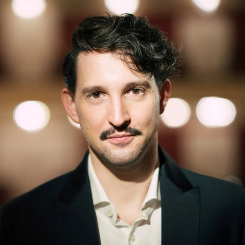 Nikolaus Habjan blickt direkt in die Kamera, im dunklen Sakko mit leichtem Lächeln – unscharfer Bühnenhintergrund, Porträt des Regisseurs und Puppenspielers.