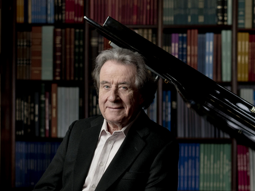 Porträt des Pianisten Rudolf Buchbinder vor einem schwarzen Flügel, im Hintergrund ein Bücherregal mit farbigen Buchrücken.