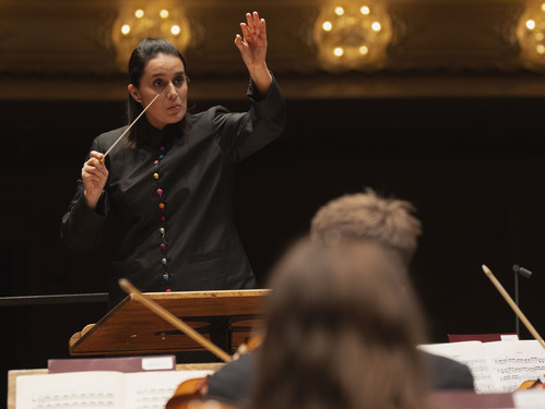 Dirigentin hebt den Taktstock vor Orchester auf beleuchteter Bühne.