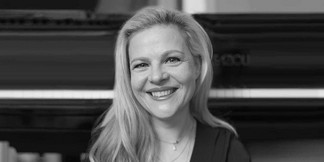 Schwarz-weiß Porträt einer lächelnden Frau (Angelika Ortner) mit langen Haaren vor einem Klavier.