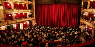 Publikum vor rotem Theatervorhang in klassisch dekoriertem Theatersaal