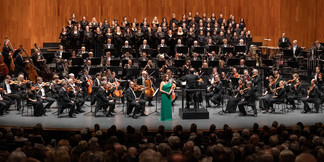 Konzert mit großem Orchester, Chor und Solistin in grünem Kleid auf Bühne vor Publikum.