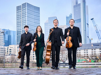 Vier Musiker:innen des Eliot Quartetts in dunkler Konzertkleidung schreiten mit ihren Streichinstrumenten selbstbewusst über das Frankfurter Mainufer, im Hintergrund moderne Hochhäuser und Baustellenkräne der Skyline.