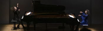 Zwei Pianistinnen spielen synchron an zwei Flügeln auf abgedunkelter Bühne vor Publikum.