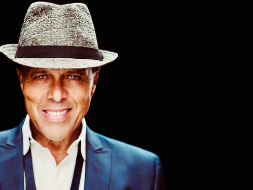 Mann mit Hut und Anzug lächelt vor schwarzem Hintergrund in die Kamera.