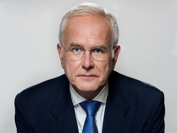 Porträt von Harald Schmidt im blauen Anzug mit blauer Krawatte vor hellem Hintergrund, frontal in die Kamera blickend.