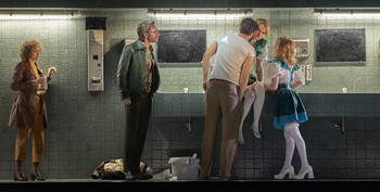 Theaterszene mit fünf Personen in gekacheltem Raum, eine Frau in blau-weißem Kleid sitzt auf Waschbecken.