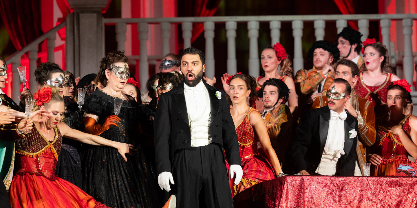 Opernszene von "La Traviata" mit Sänger im Frack und maskierten Darstellerinnen in prunkvollen Kostümen auf Bühne.