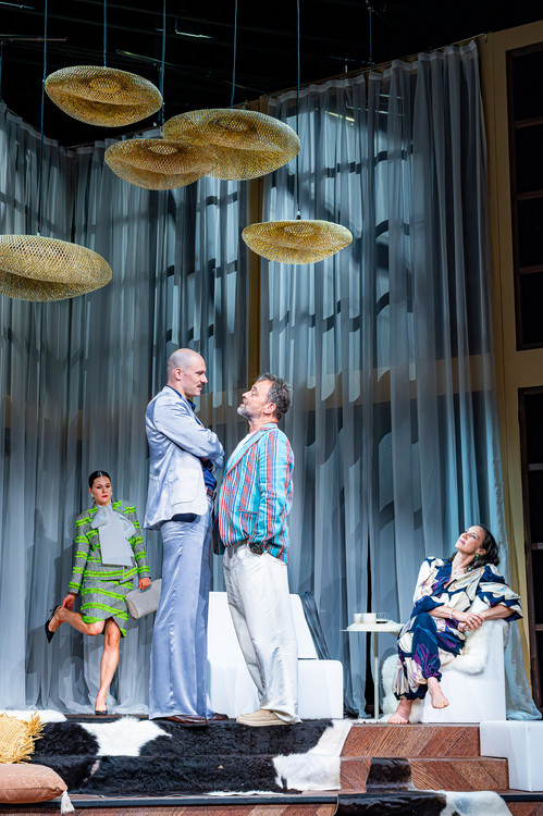 Vier Schauspieler:innen auf einer stilisierten Bühne mit modernen Möbeln und großen goldenen Deckenlampen. Zwei Männer stehen sich in einem angespannten Dialog gegenüber, während zwei Frauen im Hintergrund beobachten – eine sitzt entspannt, die andere steht mit verschränkten Armen.