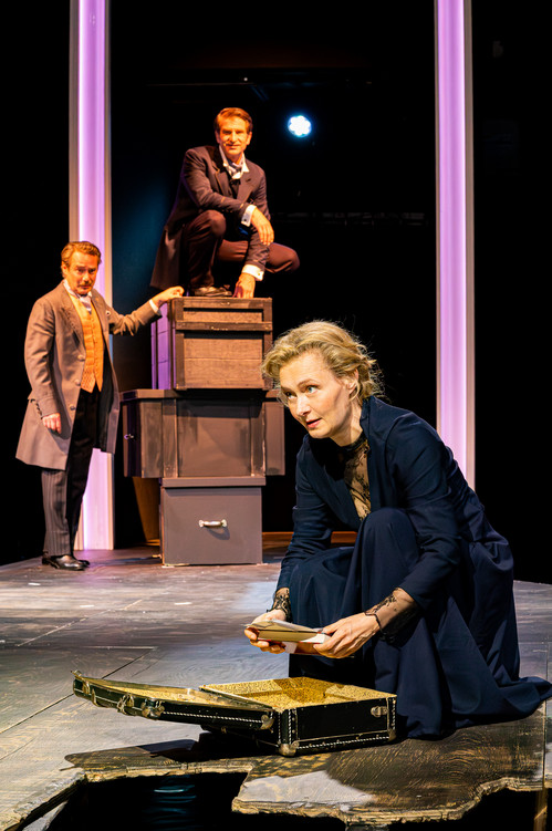 Theaterszene mit drei männlichen Schauspielern in historisch anmutenden Kostümen. Eine Frau kniet vorn am Bühnenrand, hält einen Brief in der Hand und blickt konzentriert in die Ferne. Hinter ihr stapeln sich Kisten, auf denen ein Mann sitzt, während ein weiterer daneben steht.