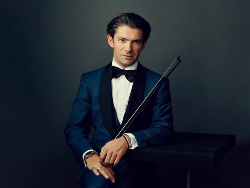 Eleganter Mann im dunkelblauen Smoking mit schwarzer Fliege, der einen Cellobogen hält, vor neutralem Hintergrund.