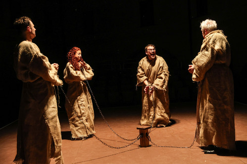 Vier Schauspieler:innen in groben, braunen Kutten stehen auf der Bühne im Kreis und sind mit Ketten aneinander gefesselt. Eine Figur schreit oder lacht wild, während die anderen sie ansehen.