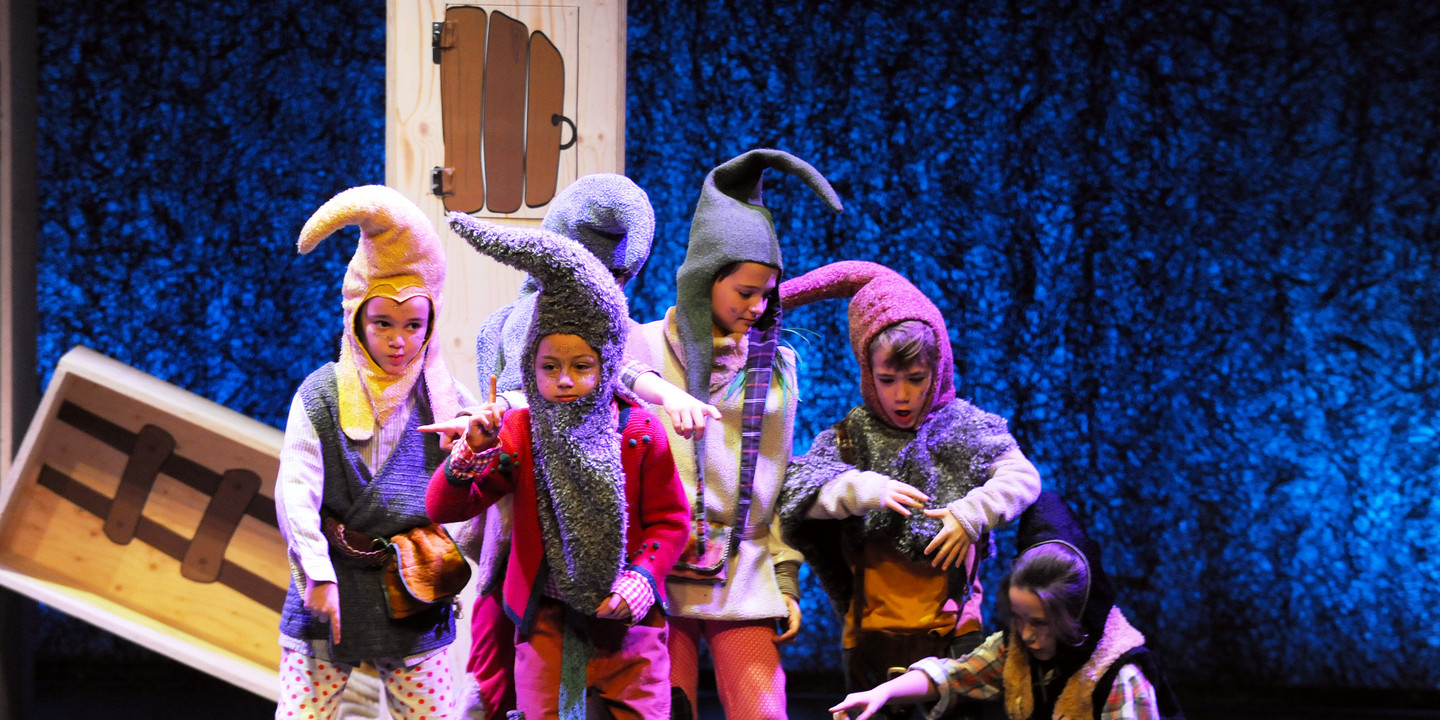 Kinder in bunten Zwergenkostümen auf einer Theaterbühne mit Requisiten und blauem Hintergrund.