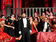 Opernszene mit Sänger im Frack und maskierten Darstellerinnen in prunkvollen Kostümen auf Bühne.