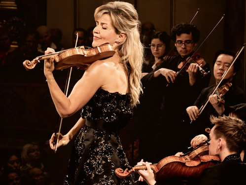 Geigerin in elegantem Kleid spielt vor Orchester auf der Bühne mit geschlossenen Augen.