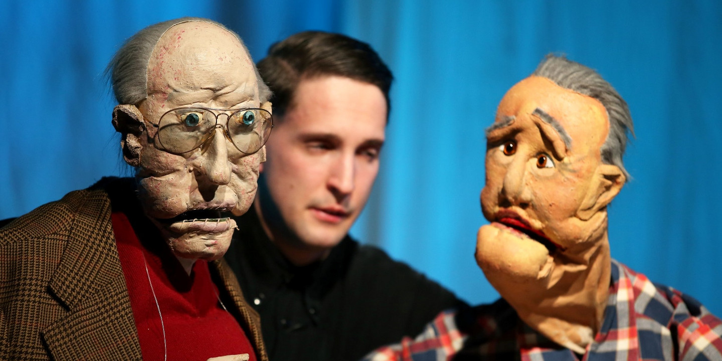 Puppenspieler mit zwei lebensechten Handpuppen auf der Bühne, die ältere Männer darstellen.