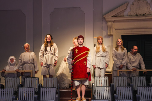 Gruppenszene in historischer Inszenierung: Mehrere Darsteller:innen in weißen Gewändern stehen in den Zuschauerrängen; im Zentrum ein junger Mann in rotem Wams mit goldenen Lilien und Stab, flankiert von weiteren Figuren in mittelalterlicher Kleidung.
