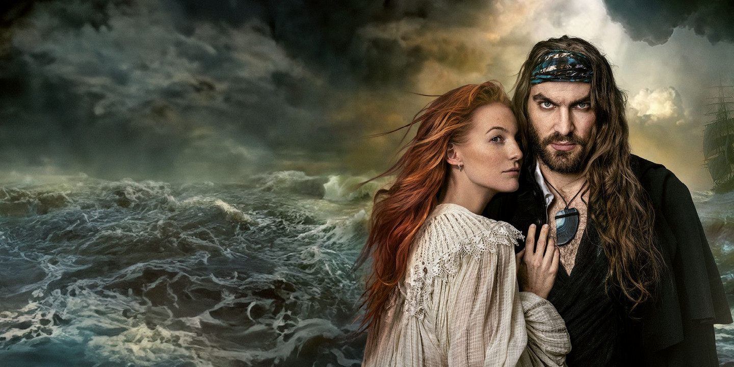 Frau mit roten Haaren und Pirat mit langem Haar stehen eng umschlungen vor stürmischer Meereskulisse.